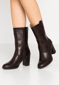 Botas de tobillo de imitación cuero marrón con acabado texturizado, cremallera lateral y tacones en bloque. Presentan una punta redondeada y costuras decorativas.