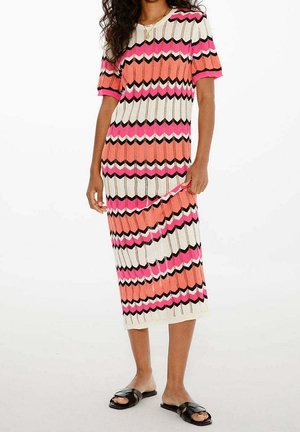 Femme portant une robe en maille mi-longue avec des rayures en zigzag roses, blanches, orange et noires, et des sandales plates noires à bout ouvert.