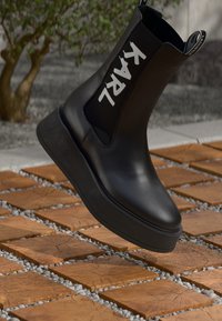 Bottines à plateforme noires avec le texte "KARL" sur le panneau latéral extensible, présentées au-dessus de carreaux en bois carrés et d'un sol en petits graviers.
