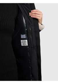 Zwarte jas met een zwarte, glanzende binnenkant, voorzien van een gestructureerd buitenoppervlak en een fleece kraag. Labels zichtbaar: "Khujo" en wasvoorschriften.