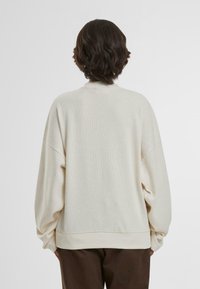 Fester, cremefarbener Strickpullover mit strukturiertem Oberflächen, lockerer Passform, überschnittenen Schultern und geripptem Saum. Die Rückansicht zeigt ein strukturiertes Design.