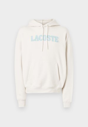 Krémová mikina s kapucí, klokáním kapsou, stahovací šňůrkou a měkkou froté texturou. Má chlupatě vyšívané logo "LACOSTE" v pastelových barvách.