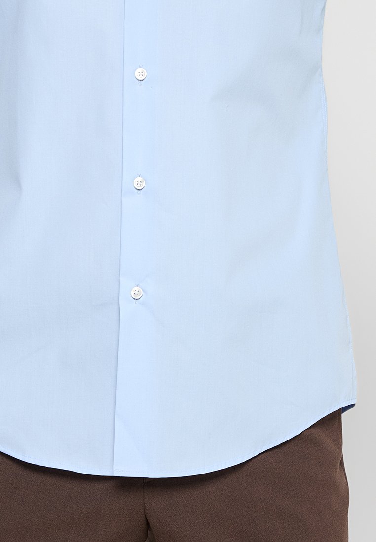 Camicia button-up azzurro chiaro infilata dentro pantaloni marroni, mostrando i dettagli della parte inferiore del torso e della sezione centrale.