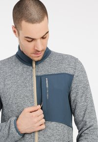 Grijze textuurjack met een blauwe paneel. Heeft een volledige rits, kinbeschermer en een borstzak met een logo. Glad en gestructureerd ontwerp.