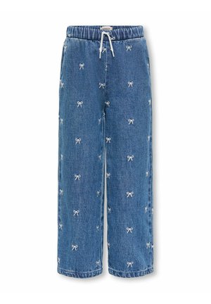 Jeune fille debout, la main sur la hanche, portant un haut rose à manches courtes, un pantalon bleu avec des motifs de nœuds blancs, et des baskets beiges.