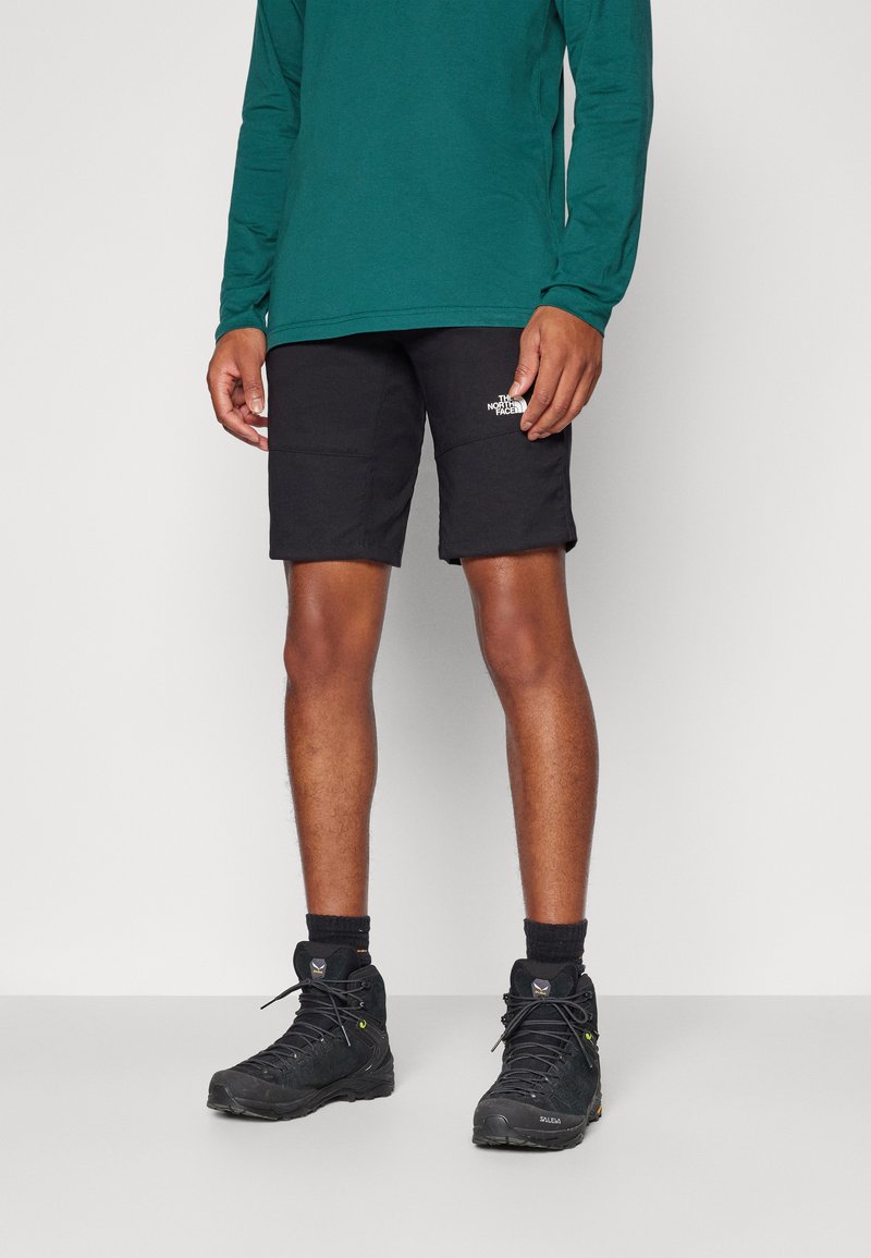 Shorts noirs au design fonctionnel, ornés d'un logo, associés à des bottes de randonnée noires et à un haut à manches longues vert.