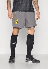 Grå sportsshorts med et gult logo, parret med sorte knæhøje sokker og sorte sportssko med et karakteristisk design.