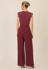 Jumpsuit borgogna con top in pizzo, maniche corte e scollatura alta. Pantaloni morbidi con spacchi laterali e chiusura con zip sul retro.