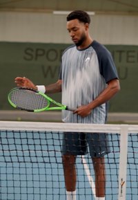 Tennis-Spieler in einem blau-grauen Verlaufsshirt und dunklen Shorts, der einen grünen Schläger hält, steht neben einem weißen Netz auf einem blauen Platz.