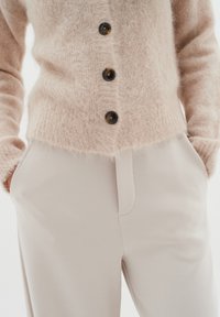 Cardigan en tricot flou rose clair avec quatre boutons foncés, associé à un pantalon beige clair avec une poche latérale et une texture lisse.