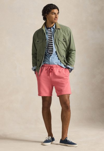 Χακί πράσινο μπουφάν με γιακά κάμελ, ριγέ μπλε ναυτικό και λευκό πουκάμισο, ανοιχτό μπλε τζιν από κάτω, κοραλί ροζ σόρτς, μπλε αθλητικά παπούτσια.