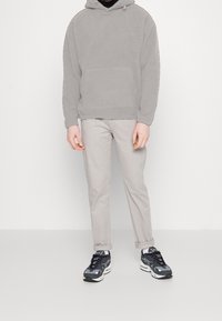 Felpa grigia in fleece con tasca a marsupio, pantaloni chino grigi chiari e sneakers nere con dettagli grigi, il tutto su uno sfondo bianco.