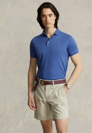 Jeune homme portant un polo bleu, un short beige avec une ceinture à rayures et une montre-bracelet, debout devant un fond neutre.