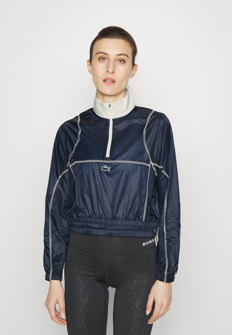 Veste de sport marine avec un col zippé à mi-hauteur, taille élastique et accents gris clair. Présente le logo Lacoste sur la poitrine, fabriquée en tissu léger.
