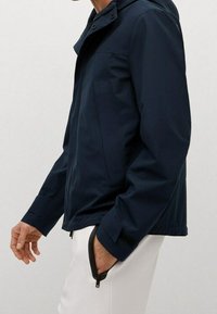 Nepromokavá námořnicky modrá bunda s texturovaným povrchem, zipovými kapsami a vysokým límcem. Má moderní, přiléhavý střih.