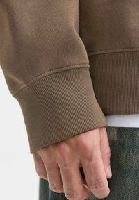Bruine sweatshirt-mouw met ribbelstructuur en een gladde afwerking. Hand die de mouw vasthoudt, waardoor de stofdetails en stiksels worden benadrukt.