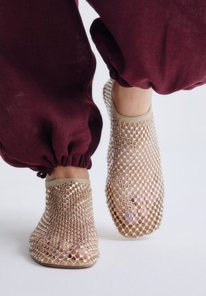 Pies que llevan zapatillas beige de malla entretejida abierta combinadas con pantalones burdeos con cordón, sobre un fondo blanco.