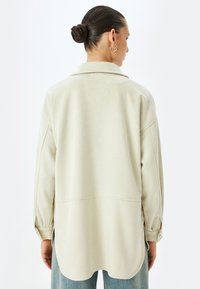 Camicia oversize beige chiaro con una texture in suede morbido, aperture laterali doppie e colletto classico. Presenta maniche lunghe e dettagli ai polsini.