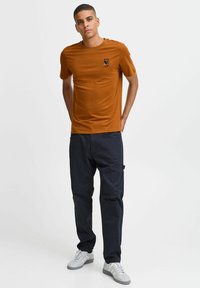 Brun crew-neck t-shirt med et lille trykt design på venstre bryst, parret med mørkeblå cargo-bukser og grå sneakers.