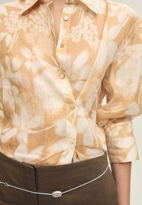 Camicia button-up beige con motivo floreale, realizzata in tessuto testurizzato e leggero. Presenta un colletto classico e una leggera lucentezza, abbinata a una vita marrone.