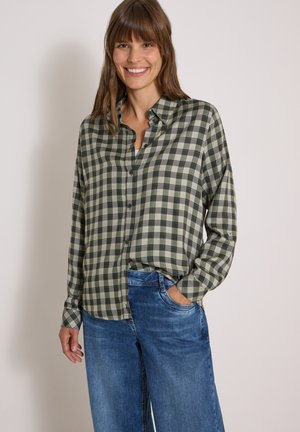 Smilende kvinde med brunt hår iført en grøn og beige ternet skjorte med knapper og blå jeans med den ene hånd i lommen.