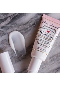 Too Faced Hangover Replenishing Face Primer in a pink tube, with a white cream texture swatched beside it on a gray surface.