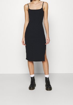 Kobieta ubrana w czarną, midi sukienkę bez rękawów z bocznym rozcięciem, białe skarpetki do kostki i czarne sznurowane botki, na tle jednolitej białej ściany.