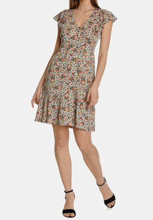 Femme portant une robe courte à fleurs avec des manches flottantes et des talons noirs à bride à la cheville, debout sur un fond clair uni.