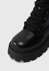 Botte en cuir noir avec semelle épaisse et rainurée et larges lacets noirs texturés noués à travers des œillets en métal.