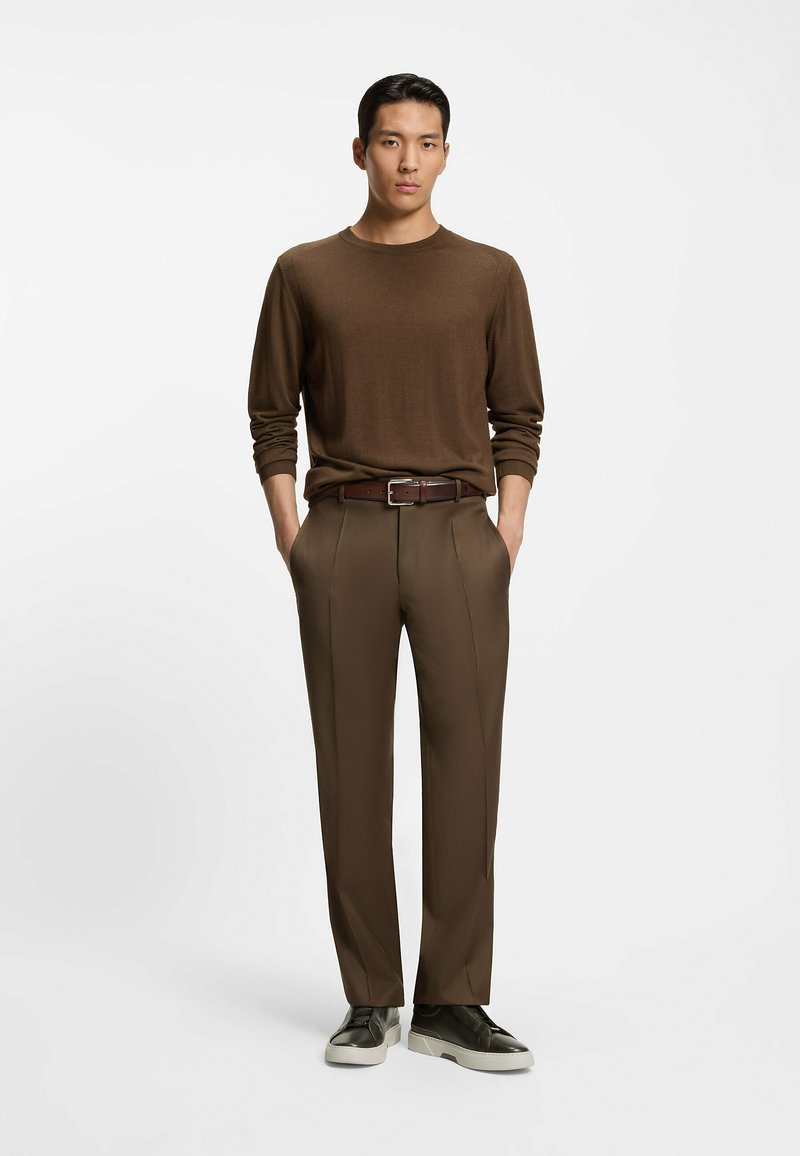 Brun långärmad tröja, skräddarsydda bruna byxor, brunt läderbälte och mörka sneakers med vita sulor, visade mot en vit bakgrund.