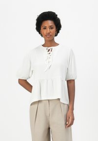 Weiße Peplum-Bluse mit Puffärmeln, verfügt über einen runden Ausschnitt und eine Schnürdetail an der Vorderseite. Kombiniert mit hellbeigen Hosen.
