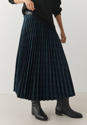 Una modelo lleva un jersey negro, una falda plisada de cuadros en tonos azul verdoso oscuro y botines negros.
