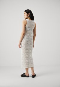 Robe débardeur blanche en crochet avec un motif de dentelle hexagonal, longueur midi, coupe ajustée. Portée avec des sandales noires, le modèle est posé de dos.