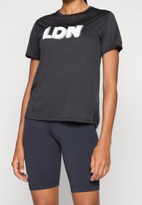 Camiseta deportiva negra con "LDN" impreso en blanco, de mangas cortas, cuello redondeado y un corte ligeramente holgado, combinada con pantalones cortos de ciclismo azul marino.