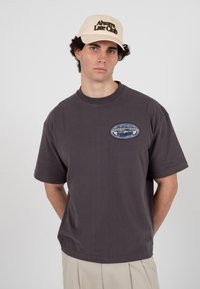 Jeune homme portant une casquette beige "Always Late Club", un t-shirt gris foncé avec un patch ovale "Prohibited Fishing Club" et un pantalon beige clair, debout immobile.