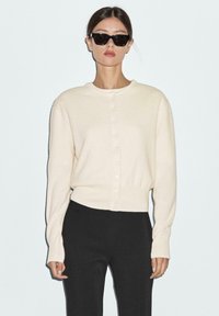 Cream knit cardigan with round neckline, front buttons, and long sleeves. Paired with black trousers. Simple, clean design.