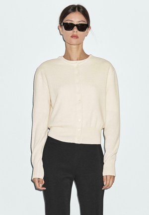 Crèmekleurige gebreide cardigan met ronde halslijn, vooraan knopen en lange mouwen. Gecombineerd met zwarte broeken. Eenvoudig, strak ontwerp.