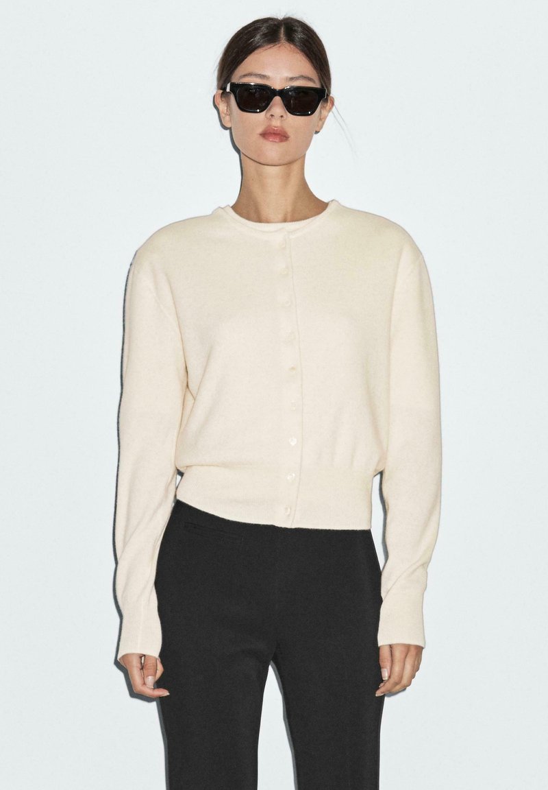 Cream knit cardigan with round neckline, front buttons, and long sleeves. Paired with black trousers. Simple, clean design.