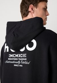 Svart hettegenser med en iøynefallende hvit logo på ryggen. Har et strukturert stoff og fôret hette. Inkluderer merkevaredetaljer i hvitt.