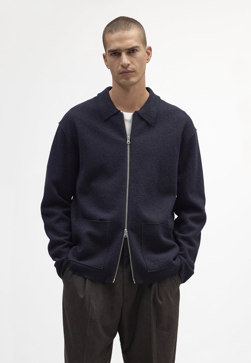 Marine blauw ritsjack gemaakt van textuurmateriaal, met een kraag, twee voorzakken en lange mouwen. Gecombineerd met donkere broeken.