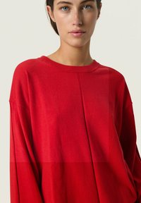Rød oversized sweatshirt lavet af blødt materiale med rund halsudskæring, sænkede skuldre og minimal syning foran.