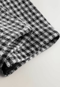 Nahaufnahme eines gefalteten Wollstoffs mit schwarz-weißer Hahnentrittmusterung, auf einer hellen Oberfläche mit sichtbarer Textur und Nähten.