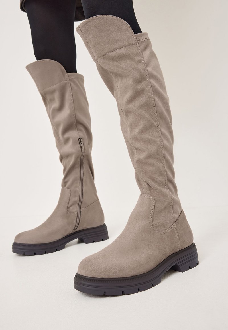 Ghete din suede bej, peste genunchi, cu un design lejer, fermoar pe lateral și talpă din cauciuc închis, cu o textură ondulată.