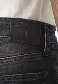 Černé denimové džíny s texturovanou koženou nášivkou označenou "TOMMY JEANS" na pásku, s kontrastním šitím a přiléhavým střihem.