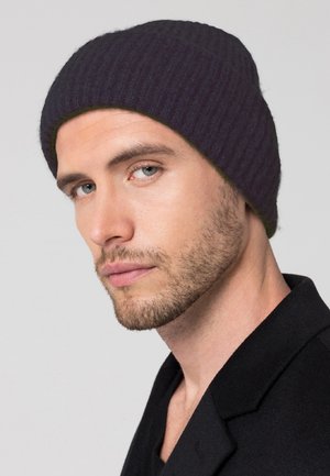 Dunkellila gerippte Strickmütze mit eng anliegendem Design, bietet eine weiche Textur und subtile Highlights. Tragen von einem Model in einem schwarzen Mantel.