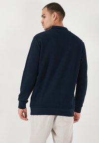 Pulover tricotat cu model rib la culoare bleumarin, cu guler, mâneci lungi și o croială lejeră. Prezintă o textură uniformă și detalii subtile la tiv.