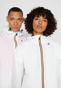 Jachete crem și roz cu fermoar și glugă, având o dungi multicolore și un logo pe pieptul stâng. Textură netedă și material ușor.