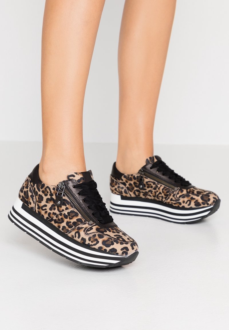 Nogi w panterkowych trampkach na platformie z czarnymi sznurowadłami, bocznym zamkiem błyskawicznym i grubymi czarno-białymi pasiastymi podeszwami na białej podłodze.