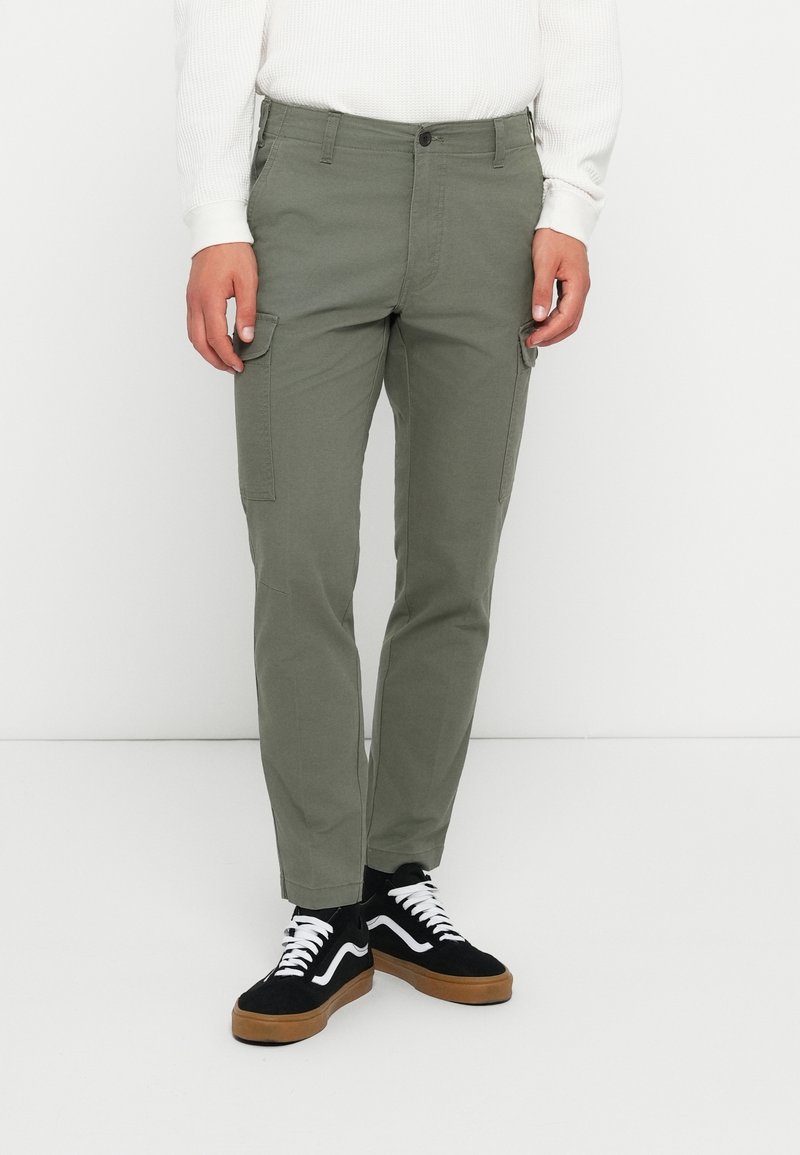 Homme portant un pantalon cargo vert olive, des baskets noires avec des lacets blancs, et un pull blanc à manches longues, debout devant un fond blanc.