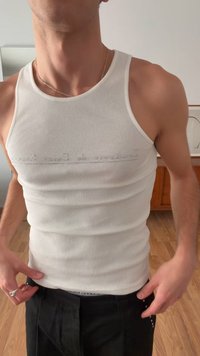 Bărbat purtând un maiou alb, conținut, cu dungi fine și text subtil pe piept, și pantaloni negri, ajustând banda de la talie în interior.